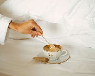 A hand delicately stirs a spoon in creamy coffee served in a china cup on a saucer, with a wrapped biscuit, placed on a bed.