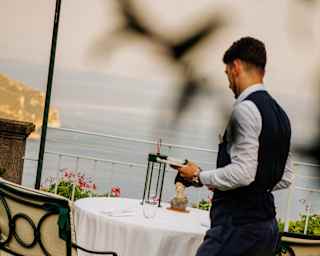 Angled image of a waiter as he prepares a white-clothed table for two on Belvedere's terrace, seen through foliage.