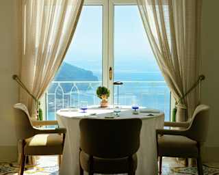 A round table with three chairs sits by glazed doors with tied-back drapes and views out on to the sunny Amalfi coast.