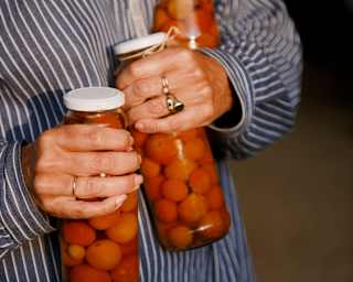 A woman in a striped shirt carries three tall jars of pickled cherry tomatoes, one in each hand and one under her left arm