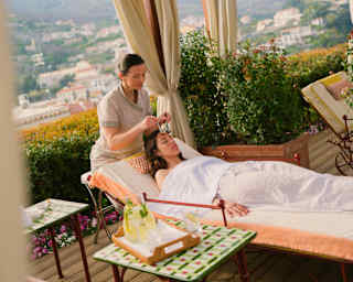 A spa therapist sits at the head of a treatment bed where she massages the forehead of a guest who lies beneath a sheet.