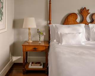 A tall bed post rises from the left side of a carved headboard belonging to a white bed with side table, in a bedroom detail.