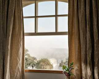 The lower half of a sash window is raised to reveal an incredible, misty falls view through a window with wheat-tone curtains.