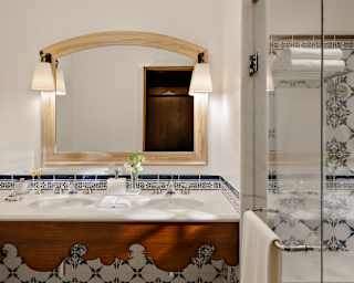 Lamps flanking a wall mirror throw light on marble twin basins on a base with a carved wood panel and blue and white tiles.