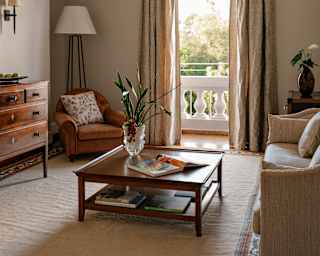A Deluxe Suite living room rests in neutral hues with a low table, sofa, chair, drawers and views out through a balcony door.