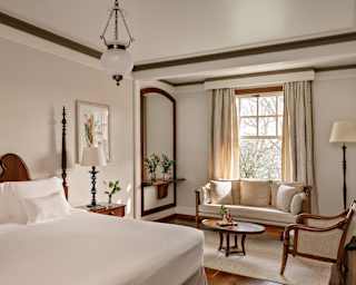 A two-seater sofa soaks in light from an open window in a Junior Suite in neutrals and wood, seen over a large white bed.