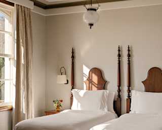 Sunlight through a window scatters across twin beds with spear-like posts and carved headboards in a Junior Suite.