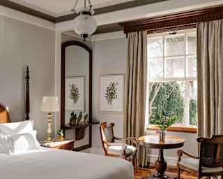 A wall mirror reflects a nature print that pairs with a window view enjoyed by table and two chairs, seen over a bed.