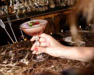 A female guest reaches for a cocktail with pink hues, soft foam and botanical garnish on the marbled bar-top at Bar Taroba.