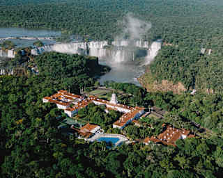 Aerial view of a resort surrounded by dense forest, with large waterfalls and mist rising in the background under a clear sky.