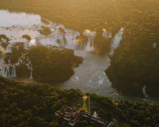Aerial view of a lush green forest with a river winding through it and several waterfalls cascading into the water, captured at sunset with golden light illuminating the landscape.