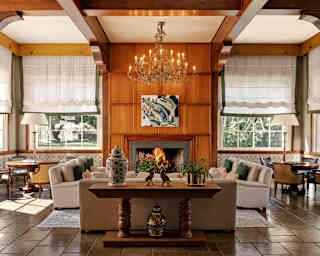In a lobby area, a candle chandelier glows above banks of armchairs by an open fire flanked by cane lounge chairs and tables.