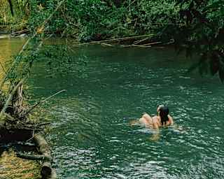 A woman in a swimming costumes sits in a shallow, emerald-green river bordered by forest, seen from behind and above.