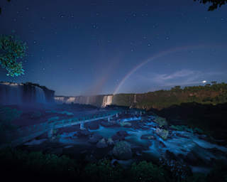 A starry night sky above Iguazu Falls, with a faint moonbow arching over the waterfalls. The river and rocks are illuminated, and a walkway crosses the foreground.