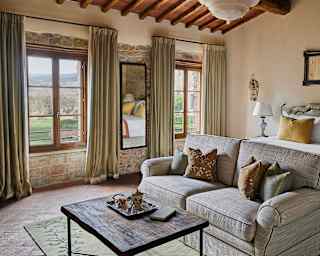 A bedroom with a sofa, bed and two big windows on to the countryside, reflects Tuscany's tones with wheat and sage fabrics.
