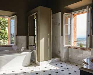 Open dual aspect windows reveal distant Tuscan hills and nearby trees. A mirror-mounted tap feeds the freestanding bathtub
