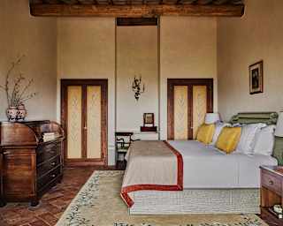 View of a bedroom with warm wood pieces and high beam ceiling over a bed with Loro Piana textiles in wheat and earth tones.