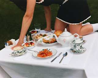 A woman in a black dress with white piping and socks crawls across a breakfast table with pastries, fruit, coffee and salmon.