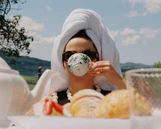 A woman with a towel hair-wrap drinks tea from a porcelain botanical-print cup, seen at eye-level over a table of pastries.