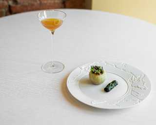 A stuffed onion and dolmades rest on a white plate with an off-centre, engraved rim, at a table with a honey-hued cocktail.