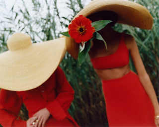 Two women in red, one in a dress, the other in skirt and crop top, wear huge straw hats, obscuring their faces in the garden.