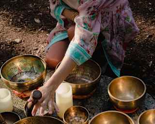 A woman in a pink and blue floral gown sits by a collection of singing bowls, varying in size, at a sound healing session.