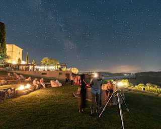 Some guests sit on the amphitheatre steps with lamps, while others stand by a telescope below a night sky sprayed with stars.