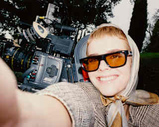 A blonde woman with a headscarf and orange sunglasses smiles in a selfie that captures a sophisticated movie camera behind.