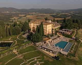 Aerial view of Castello di Casole atop a hill, set in gardens with a large pool, dining terraces and ancient lawn amphitheatre.