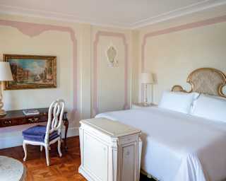 A refined bedroom with parquet floors, painting of 19th Century Venice, gilt-edged headboard and impossibly crisp sheets