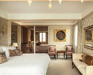A gilt framed Old Master hangs on the ivory patterned wall of a majestic bedroom with baroque furniture and kingsize bed