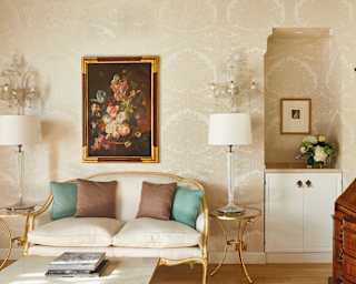 A gilt-edged cream sofa with brown and turquoise cushions sits centre below a framed still life of flowers in oils