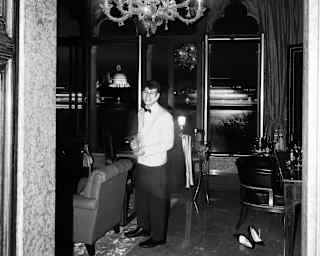 A waiter in a white jacket smiles to the camera as he serves wine in a private lounge below a Murano glass chandelier