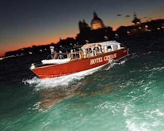 The Hotel Cipriani private launch cuts a bow wave through the green water at dusk. In the background, the lights of St Marks