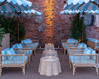 Two rows of three cane and blue chairs face each other over candle-lit rattan tables in a brick-walled bar area, seen at dusk.