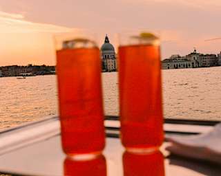 Two tall orange drinks sit on a tray in soft-focus foreground, in a view over a canal, waters peach beneath a sunset sky.