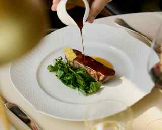 A waiter drizzles rich gravy from a small jug onto a dish of grilled lamb loin with steamed wild chicory greens at Oro.