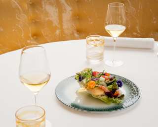 A Seafood Caesar Salad topped with edible flowers sits on a table between two place settings with white wine and water, at Oro.