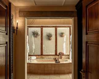 Three plants hang from a wood-framed glass splashback above a marble-tiled bath in a view through open bathroom doors.