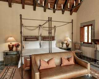 A leather sofa abuts adjacent twin four-poster beds in a cool room with a rug, stone flooring, fireplace and beamed ceiling.