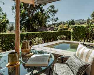 View across a sunny Valley View terrace with a raised square plunge pool, seen over table with a book and a cushioned chair.