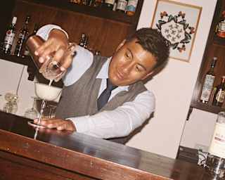 A barman in a waistcoat and tie pours a cocktail from a shaker into a coupe glass behind a wooden bar.