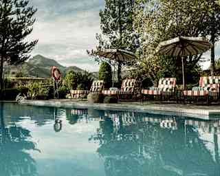 A serene outdoor swimming pool with loungers and parasols beside it, surrounded by tall trees and greenery, with mountains visible in the background under a partly cloudy sky.