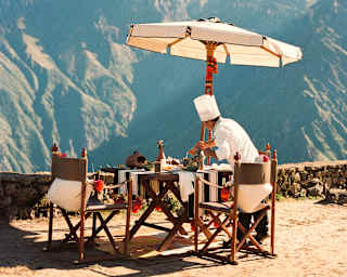 A dining table with chairs and a large parasol is set up outdoors on a mountaintop, overlooking scenic mountains under a partly cloudy sky. The table is decorated for a meal, creating a picturesque setting.