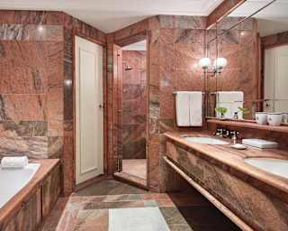 Pink marble tiles cover every surface of a large bathroom with tub, walk-in shower, double vanity and enormous mirror