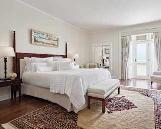 A large bedroom with views of the sea features a white topped king bed, white armchairs and antique rug on a polished floor