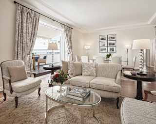 A large airy room with doors open onto a balcony is furnished with a cream sofa, armchairs and dining table that seats eight