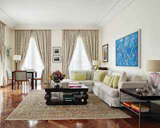 Full height windows flood a sitting room with light. A blue abstract painting hangs over a large, soft L-shaped sofa
