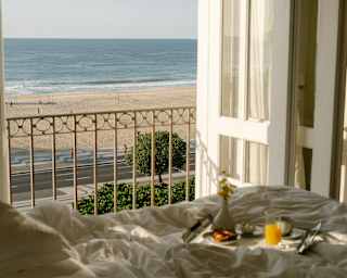 A breakfast tray rests in a shaft of sunlight on a bed with views through open doors and balcony railings of the empty beach.