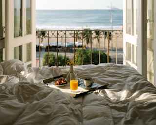 A breakfast tray with juice, coffee and pastries rests on a rumpled bed next to Juliette balcony doors, open to beach views.